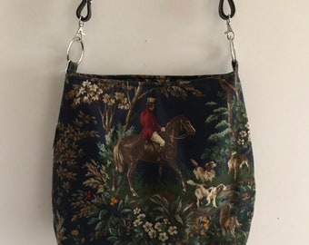 May include: A navy blue crossbody bag with a hunting scene print. The bag features a rider on horseback, dogs, and foliage. It has a braided strap and silver-tone hardware.