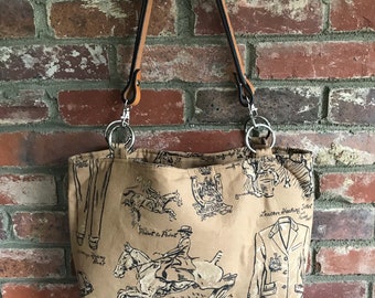 May include: Tan tote bag with black equestrian-themed illustrations, including riders on horses, riding attire, and text. The bag has brown leather straps and silver metal rings. The background is a red brick wall.