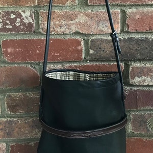 May include: A black leather tote bag with a brown leather strap and a plaid lining. The bag is hanging from a hook on a brick wall.