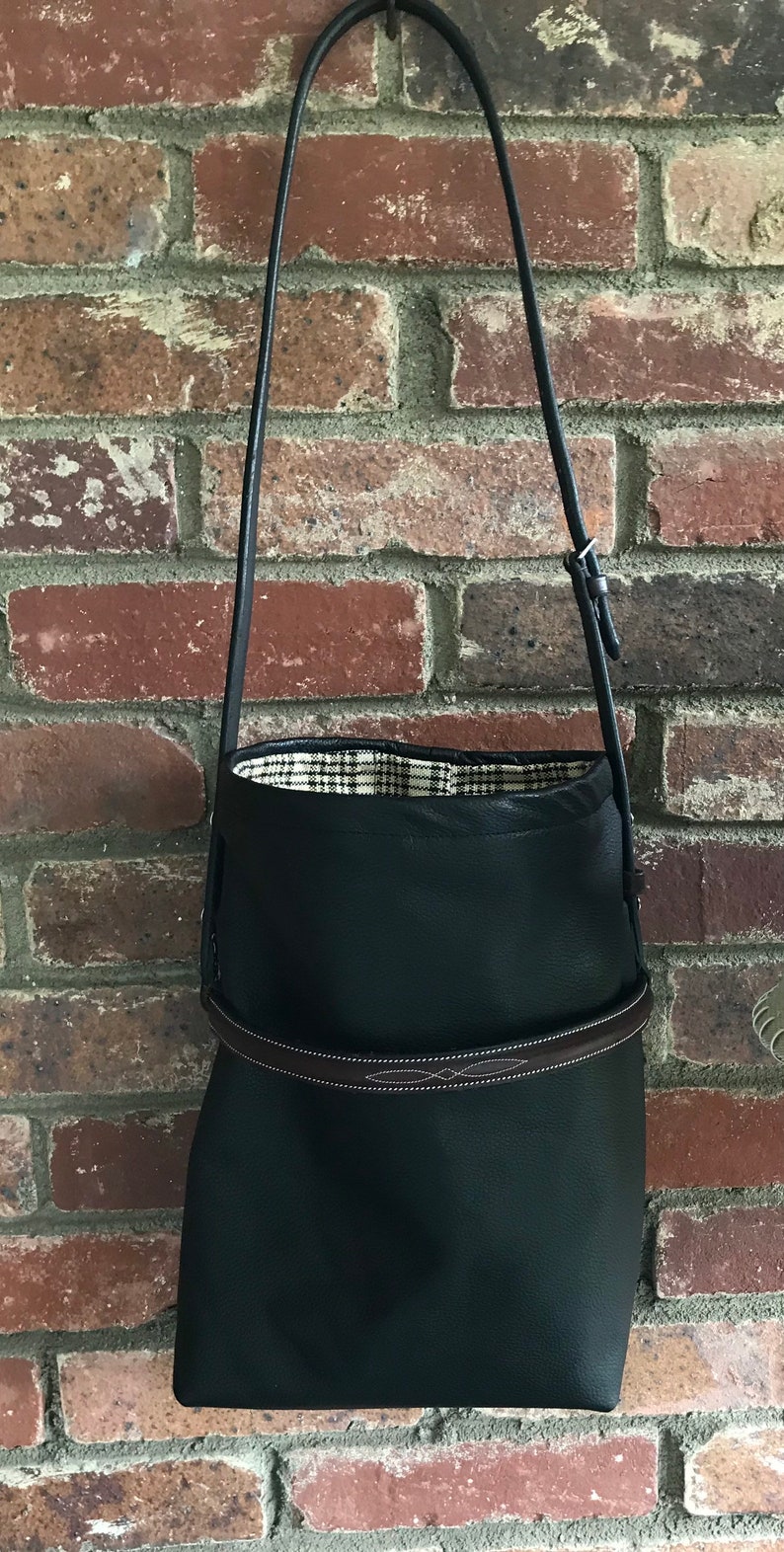 May include: A black leather tote bag with a brown leather strap and a plaid lining. The bag is hanging from a hook on a brick wall.
