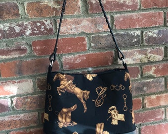 May include: A black leather tote bag with a brown and black equestrian print. The bag has a long, braided strap and a black leather bottom.