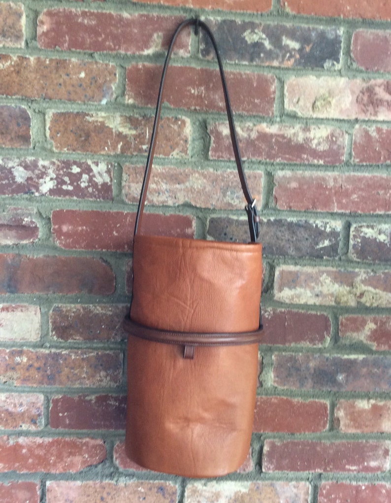 May include: A brown leather bucket bag with a black strap. The bag has a brown leather trim around the top.