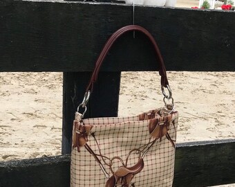 May include: A brown and white plaid handbag with a brown leather strap and silver hardware. The bag has a saddle and bridle design.