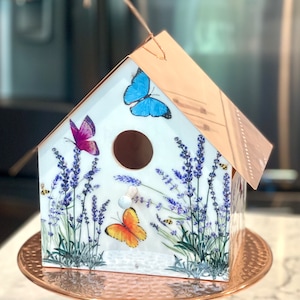 May include: A decorative birdhouse with a copper-colored roof and base. The white birdhouse features a lavender and butterfly design, with a round entry hole. The birdhouse is displayed on a hammered copper-colored pedestal.