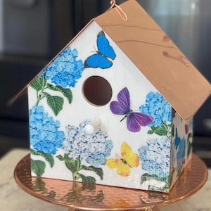May include: Decorative birdhouse with a copper-colored roof and base. The white birdhouse is adorned with blue hydrangea flowers and colorful butterfly decals. A small, matching birdhouse is also present.