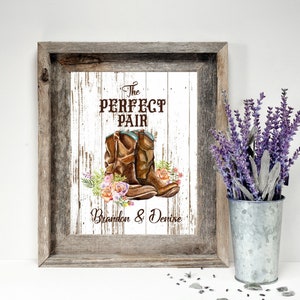 May include: Framed artwork featuring a watercolor illustration of brown cowboy boots with floral accents. The text "The Perfect Pair" is above the boots, with the names "Brandon & Denise" below. The frame is rustic wood.
