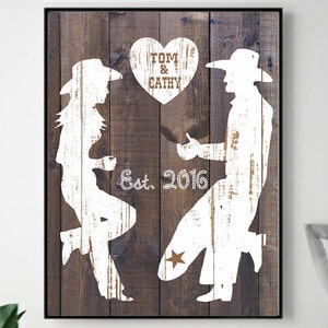 May include: Rustic wood-styled canvas print featuring white silhouettes of a couple in cowboy hats. The man is proposing to the woman.  The text 'Tom & Cathy Est. 2016' is displayed with a heart shape above the couple.  The artwork is a romantic, country-themed wedding or anniversary gift.