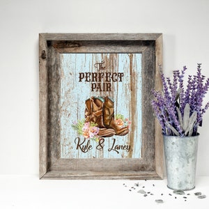 May include: Framed rustic artwork with a light blue and brown background. The artwork features a pair of brown cowboy boots with floral accents and the text "The Perfect Pair" and names "Kyle & Laney". A metal bucket with lavender is to the right.