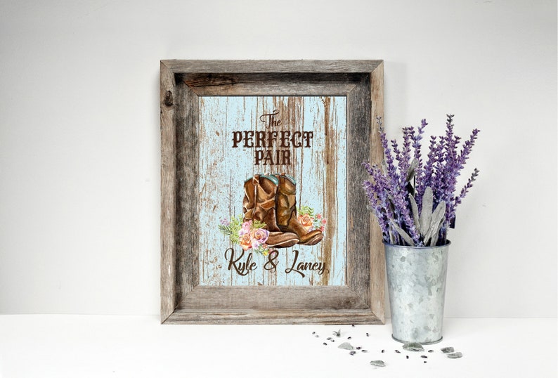 May include: Framed rustic artwork with a light blue and brown background. The artwork features a pair of brown cowboy boots with floral accents and the text "The Perfect Pair" and names "Kyle & Laney". A metal bucket with lavender is to the right.
