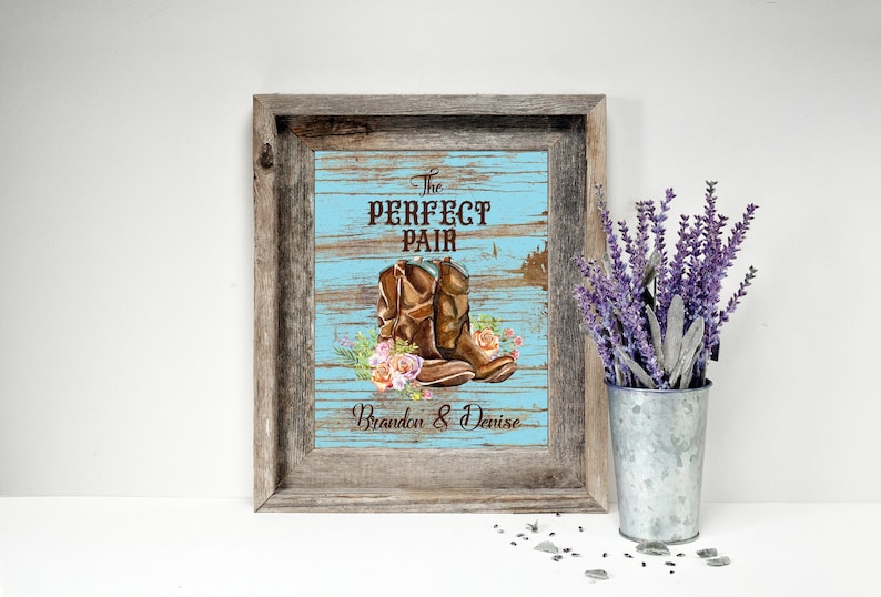 May include: Framed artwork featuring brown cowboy boots with floral accents on a blue wooden background. The text "The Perfect Pair" and names are displayed. The frame is rustic wood. A vase of lavender is next to the frame.