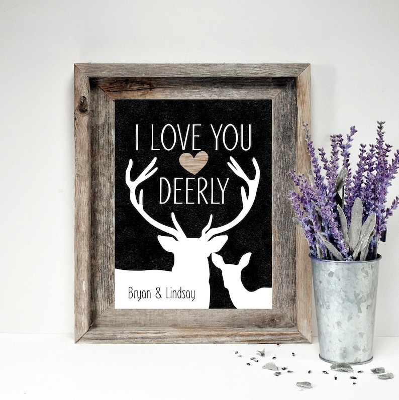 May include: A black and white print with a deer silhouette and the text "I love you deerly" with a heart in the middle of the word "deerly". The print is framed in a rustic wooden frame and has the names "Bryan & Lindsay" at the bottom.