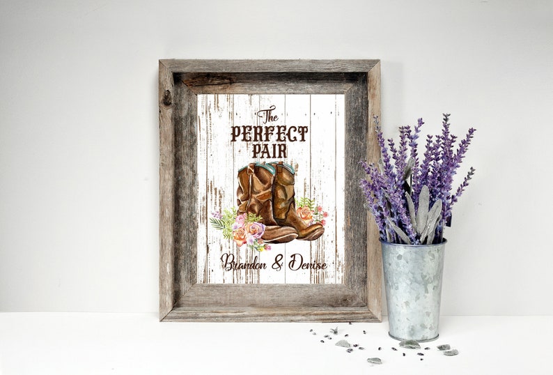 May include: Framed artwork featuring a watercolor illustration of brown cowboy boots with floral accents. The text "The Perfect Pair" is above the boots, with the names "Brandon & Denise" below. The frame is rustic wood.