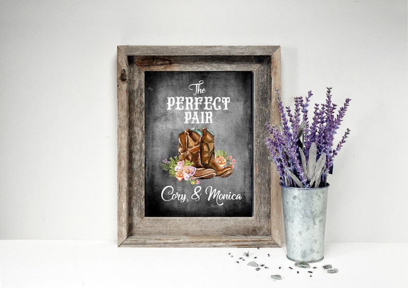 May include: Framed rustic artwork with the text "The Perfect Pair" above a watercolor illustration of cowboy boots and flowers. The names "Cory, & Monica" are below the boots. A galvanized metal vase holds lavender.