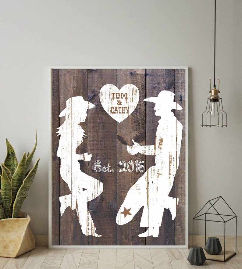 May include: White silhouette of a couple in cowboy hats, with the woman on one knee and the man standing, on a rustic wood background. The text "TOM & CATHY" is in a heart shape above them, and "Est. 2016" is below them.