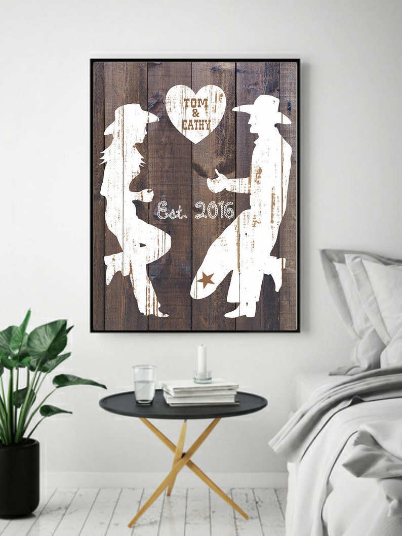 May include: Rustic wood-styled canvas print featuring white silhouettes of a couple in cowboy hats. The man is proposing to the woman.  The text 'Tom & Cathy Est. 2016' is displayed with a heart shape above the couple.  The artwork is a romantic, country-themed wedding or anniversary gift.