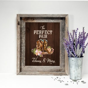 May include: A framed print with a rustic wooden frame. The artwork features a pair of brown cowboy boots with floral accents and the text "The PERFECT PAIR" and "Johnny, & Mary". A galvanized metal bucket holds lavender.