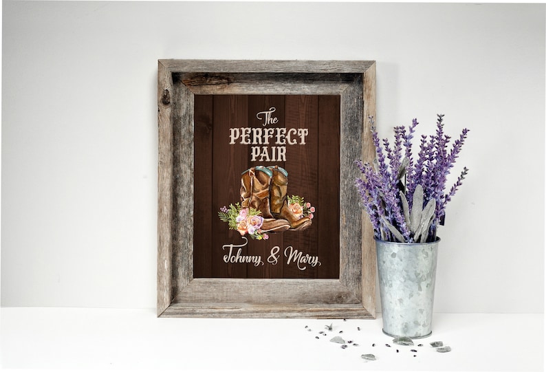 May include: A framed print with a rustic wooden frame. The artwork features a pair of brown cowboy boots with floral accents and the text "The PERFECT PAIR" and "Johnny, & Mary". A galvanized metal bucket holds lavender.