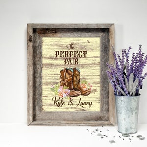 May include: A framed print with a rustic wooden frame. The print features brown cowboy boots with floral accents and the text "The Perfect Pair" above the boots and "Kyle & Laney" below. A metal bucket with purple flowers is to the right.