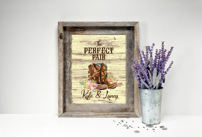May include: A framed print with a rustic wooden frame. The print features brown cowboy boots with floral accents and the text "The Perfect Pair" above the boots and "Kyle & Laney" below. A metal bucket with purple flowers is to the right.