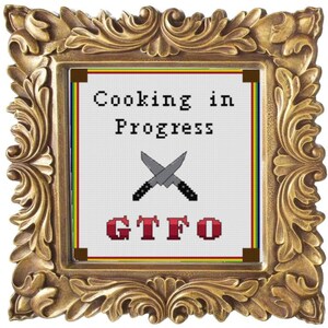 Op de afbeelding: Een ingelijst kruissteekontwerp met de tekst "Cooking in Progress" en "GTFO" in rode letters. Twee gekruiste keukemessen zijn afgebeeld in het midden van het ontwerp.