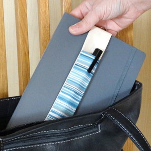 May include: A gray notebook with a striped pen holder and a black pen is being placed into a black leather bag. The pen holder has blue and white stripes. The notebook is held by a person.