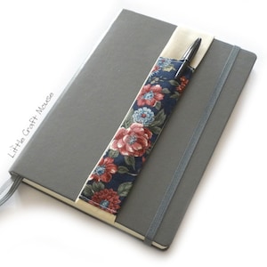 Può includere: Un quaderno grigio con un portapenne in tessuto floreale attaccato alla copertina. Il portapenne è realizzato in tessuto crema con un motivo floreale in tonalità di rosso, blu e verde. Il quaderno ha una chiusura elastica grigia.
