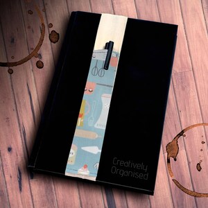 May include: A black notebook with the text Creatively Organised embossed on the cover. A vertical fabric band features a light blue pattern of kitchen utensils and baked goods, with a black pen tucked into a loop on the band.