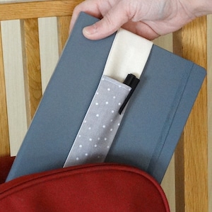 May include: A gray notebook with a white polka dot pen holder attached. The pen holder has a white fabric strap and a black pen is inserted.