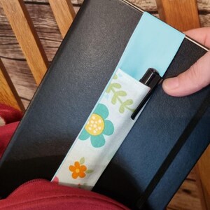May include: A black notebook with a blue and white fabric pen holder with a floral pattern. A black pen is inserted into the holder.