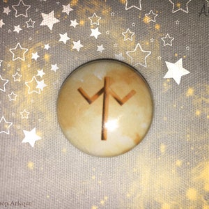 May include: A round, beige-colored button with a brown rune symbol. The button has a marbled effect and is set against a gray background with white star outlines and golden sparkles. The Etsy shop name "Arleque" is visible.