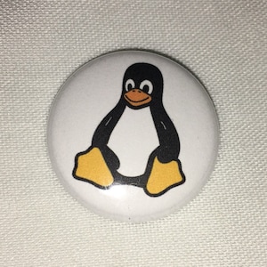 May include: A white button with a cartoon penguin illustration. The penguin is black with a yellow beak and feet. The button is round and sits on a white fabric surface.