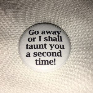 Peut inclure: Un bouton blanc avec le texte noir : "Go away or I shall taunt you a second time!". Le bouton rond est posé sur un tissu blanc.