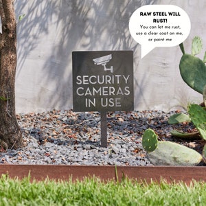May include: A black metal sign with the text "SECURITY CAMERAS IN USE" and a silhouette of a security camera. The sign is mounted on a metal stake and is placed in a bed of gravel. A speech bubble above the sign reads "RAW STEEL WILL RUST! You can let me rust, use a clear coat on me, or paint me."