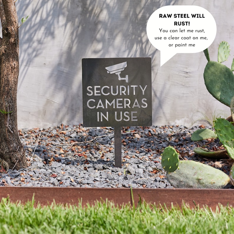 May include: A black metal sign with the text "SECURITY CAMERAS IN USE" and a silhouette of a security camera. The sign is mounted on a metal stake and is placed in a bed of gravel. A speech bubble above the sign reads "RAW STEEL WILL RUST! You can let me rust, use a clear coat on me, or paint me."