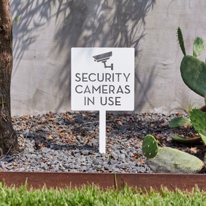 May include: White sign with a black security camera icon and the text "SECURITY CAMERAS IN USE". The sign is mounted on a white stake and is placed in a bed of gravel.