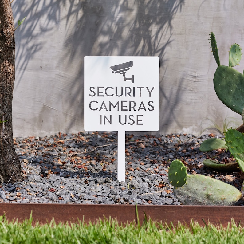 May include: White sign with a black security camera icon and the text "SECURITY CAMERAS IN USE". The sign is mounted on a white stake and is placed in a bed of gravel.