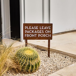 May include: A rusty brown metal sign with white text that reads "PLEASE LEAVE PACKAGES ON FRONT PORCH". The sign is on a wooden stake and is placed in a bed of gravel with a cactus plant in the foreground.