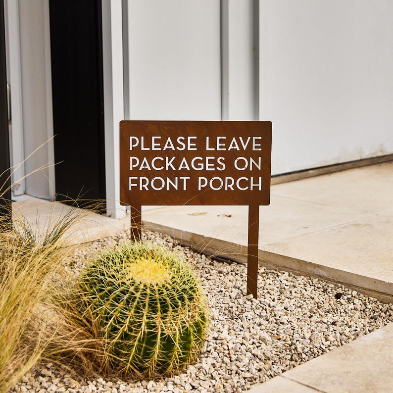 May include: A rusty brown metal sign with white text that reads "PLEASE LEAVE PACKAGES ON FRONT PORCH". The sign is on a wooden stake and is placed in a bed of gravel with a cactus plant in the foreground.