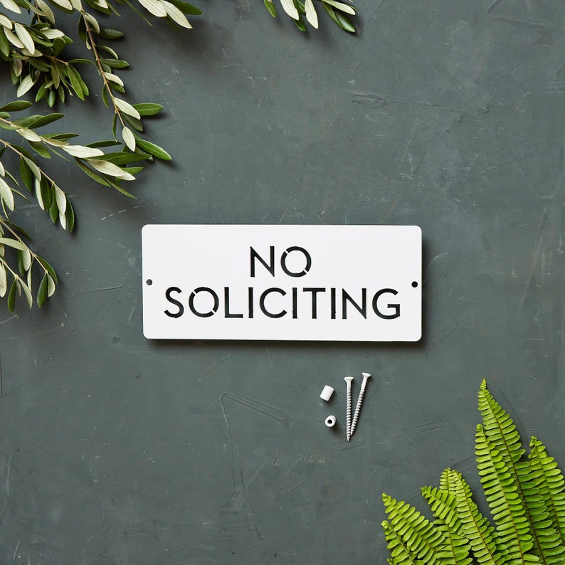 Op de afbeelding: Een wit metalen bord met de tekst "NO SOLICITING" in zwarte letters. Het bord is op een grijze achtergrond met groen gebladerte aan beide kanten.