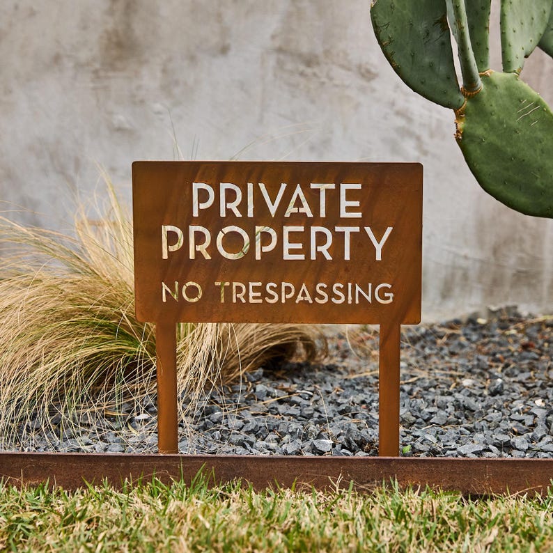 May include: A rusty metal sign with white lettering that reads "PRIVATE PROPERTY NO TRESPASSING". The sign is mounted on two metal stakes and is placed in a bed of gravel.