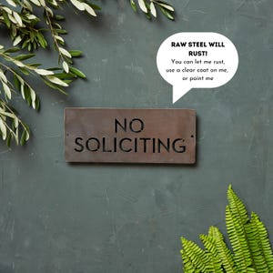 Op de afbeelding: Een rustiek metalen bord met de woorden "NO SOLICITING" in een uitgesneden ontwerp. Het bord is gemaakt van ruw staal en heeft een verweerde, roestige uitstraling. Een spraakballon boven het bord luidt "RAW STEEL WILL RUST! You can let me rust, use a clear coat on me, or paint me."