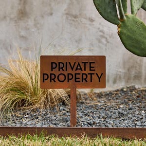 May include: A rectangular metal sign with a rusted finish featuring the text PRIVATE PROPERTY in bold black capital letters. The sign is mounted on a thin metal stake and is positioned upright in a garden setting with dry grass and gravel.