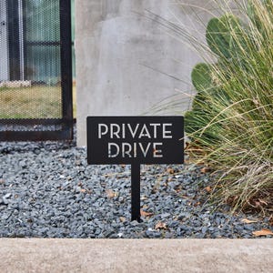 May include: Black metal sign with the words "PRIVATE DRIVE" in white lettering. The sign is mounted on a black metal stake and is placed in a bed of gravel.