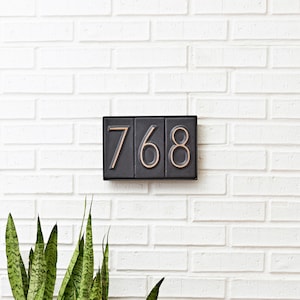May include: Black house number plaque with white numbers 768 on a white brick wall.