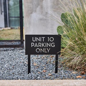 May include: Black metal sign with white text that reads "UNIT 10 PARKING ONLY". The sign is on two black metal stakes and is in a gravel area.