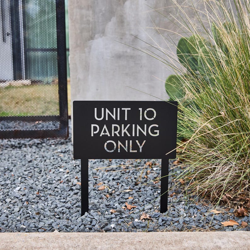 May include: Black metal sign with white text that reads "UNIT 10 PARKING ONLY". The sign is on two black metal stakes and is in a gravel area.