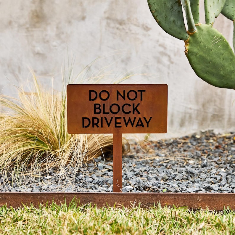 May include: A rectangular, rust-colored metal sign on a stake featuring the black text DO NOT BLOCK DRIVEWAY. The sign is positioned upright in a garden setting with gravel, dry ornamental grasses, and a green cactus visible in the background.
