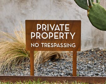 May include: A rectangular, rust-colored metal sign featuring the cut-out text "PRIVATE PROPERTY" on the top two lines and "NO TRESPASSING" on the bottom line. The sign is supported by two vertical metal stakes extending from the bottom edge.