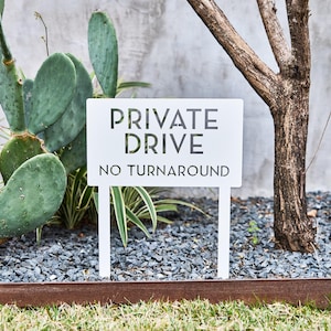 May include: White metal sign with black text that reads "PRIVATE DRIVE NO TURNAROUND". The sign is mounted on two white metal stakes and is placed in a gravel garden bed.