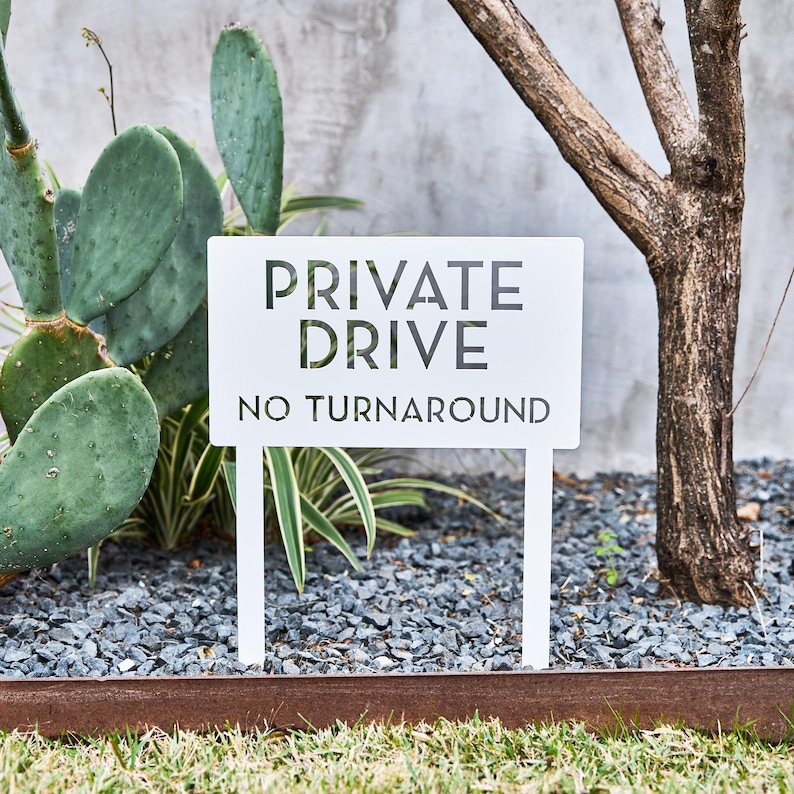 May include: White metal sign with black text that reads "PRIVATE DRIVE NO TURNAROUND". The sign is mounted on two white metal stakes and is placed in a gravel garden bed.